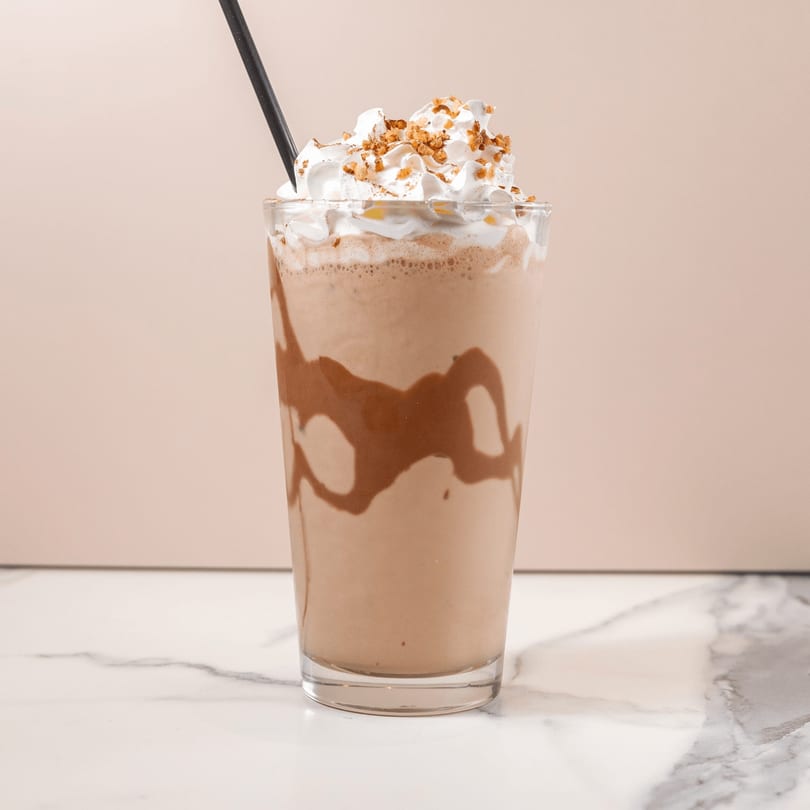 Chocolate milkshake with whipped cream, nuts, and a black straw on a marble counter.