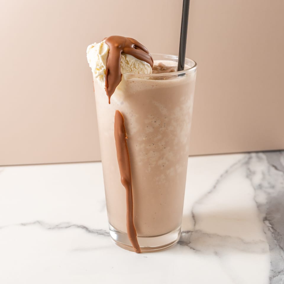 Chocolate milkshake with a scoop of vanilla ice cream and chocolate syrup drizzle, on a marble surface.