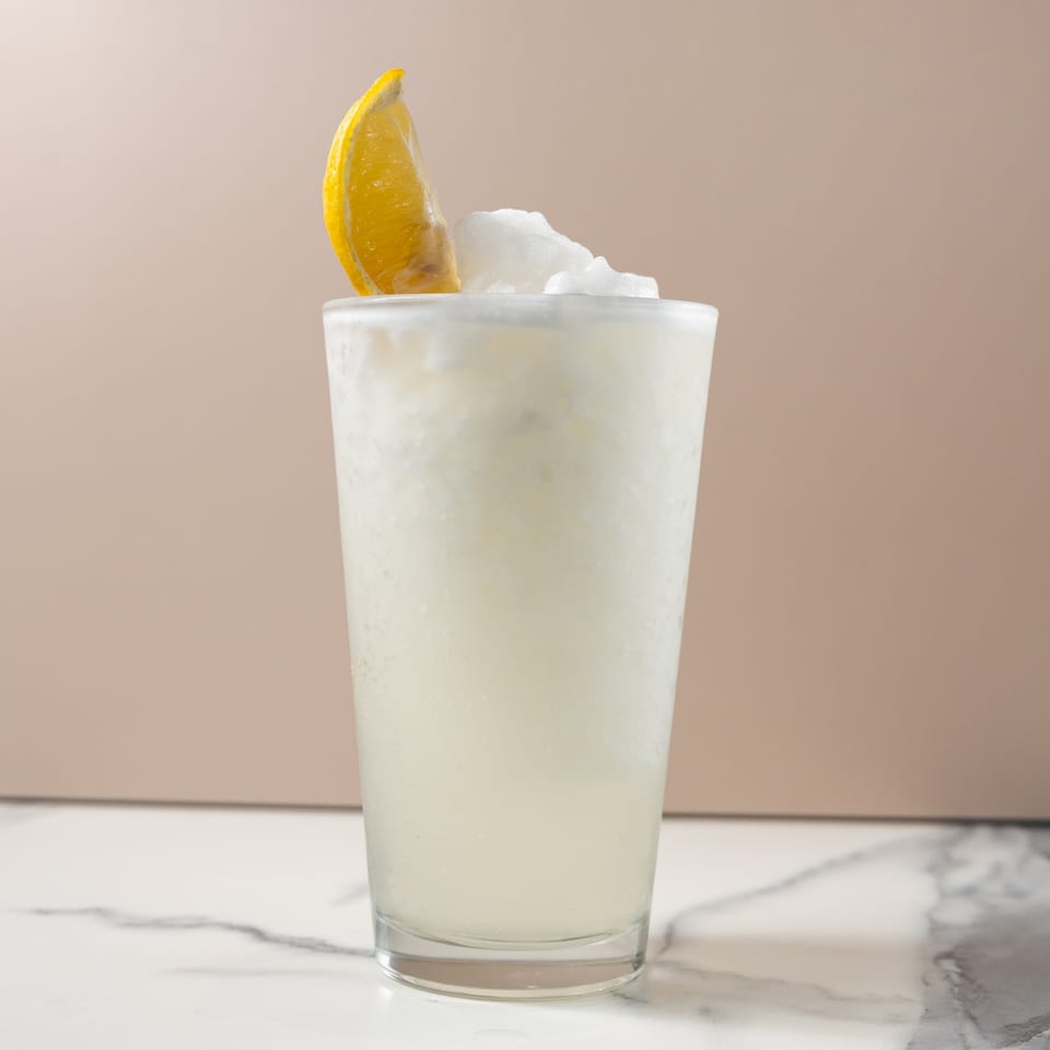 A tall glass of frozen lemonade, garnished with a lemon wedge and white shaved ice.