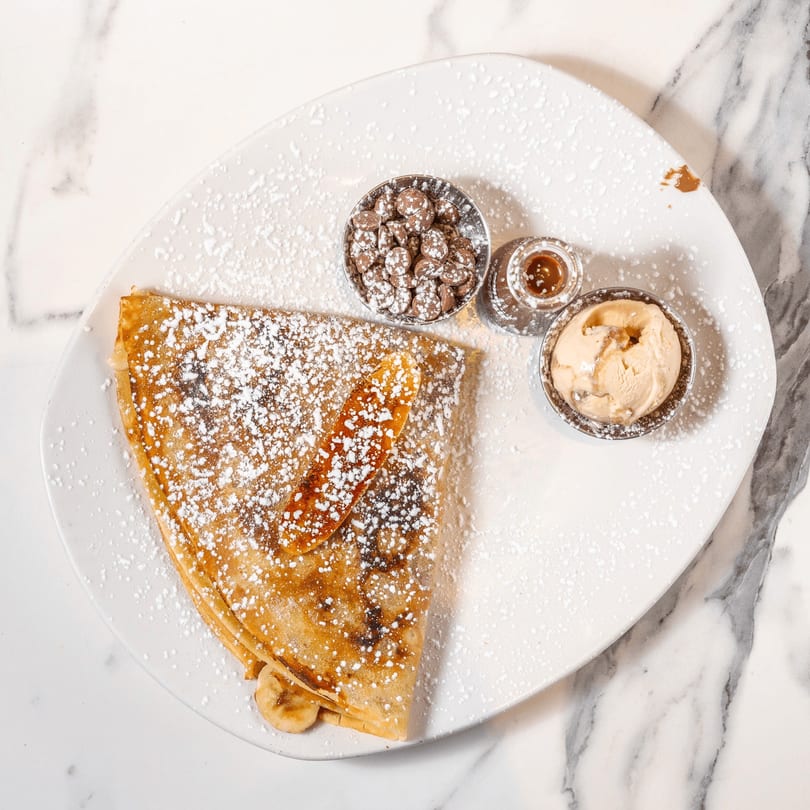 A banana and chocolate crepe dusted with powdered sugar, served with chocolate chips, caramel, and ice cream.