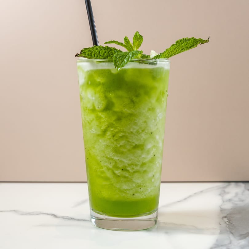 Green smoothie with mint leaves and a black straw in a tall glass on a marble surface.