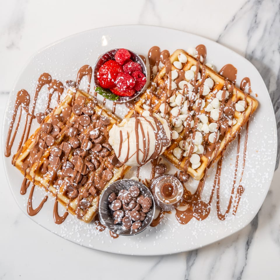 Two waffles with chocolate chips, white chocolate, ice cream, raspberries, and chocolate drizzle.