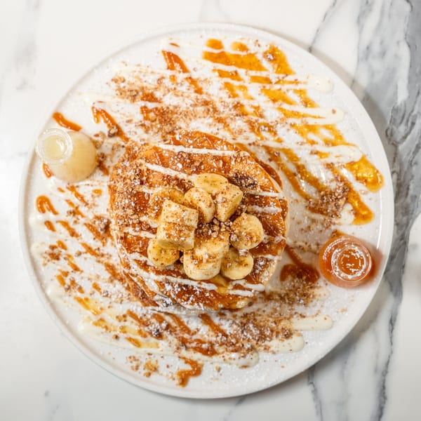 Overhead view of banana pancakes with syrup, powdered sugar, and nuts on a white plate.