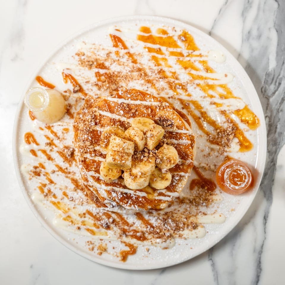 Overhead view of banana pancakes with syrup, powdered sugar, and nuts on a white plate.
