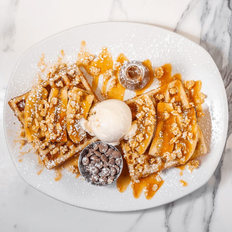 Waffles topped with caramelized bananas, ice cream, chocolate chips, caramel sauce, and powdered sugar.