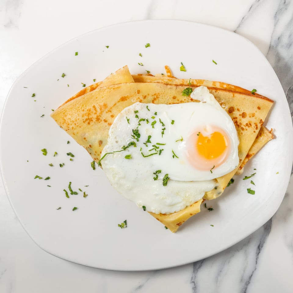 Savory crepe topped with a fried egg, melted cheese, and fresh parsley on a white plate.