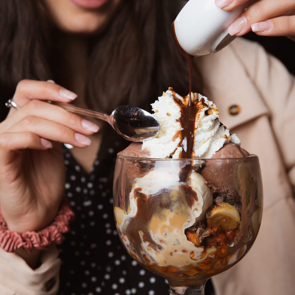 A person pouring chocolate sauce over a large Max Brenner ice cream sundae with whipped cream.